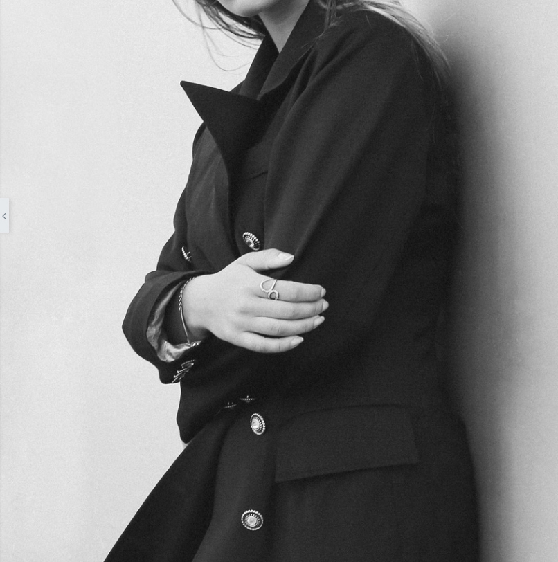 Schwarz-Weiß-Foto von weiblichem Model, sichtbar vom Kinn bis zur Hüfte, das gehobene Fashion und einen Ring und einen Armreif trägt – mit verschränkten Armen lässig an eine Wand lehnend 